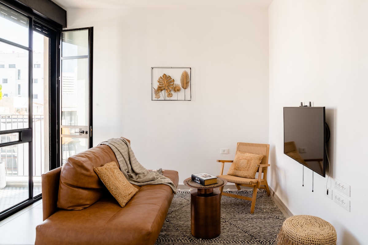 This inviting living space includes a brown leather sofa positioned near a sleek flat-screen TV. A decorative rug spans the floor, complemented by a round coffee table and a light wooden armchair. Natural light is brought in through the large glass doors, enhancing the open layout.