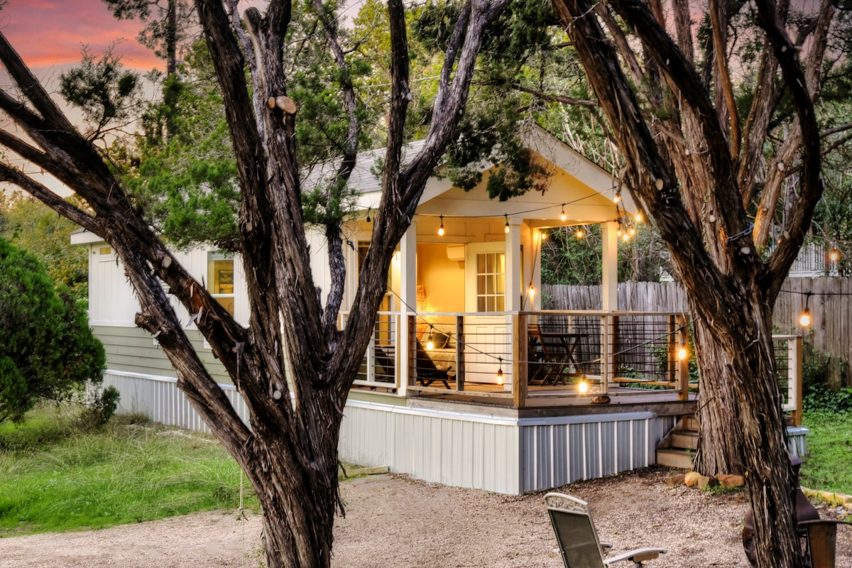 The Tiny Home is framed by tall trees, showcasing a welcoming deck lit by warm string lights. The exterior features light-toned siding, and large windows provide glimpses of the cozy interior. An outdoor seating area is visible, surrounded by natural greenery.