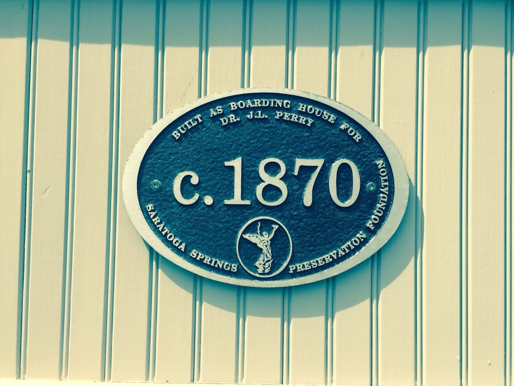A historic plaque is displayed on a white wooden wall, indicating the building was constructed around 1870 as a boarding house for Dr. J. Perry. The plaque features a circular design with contrasting dark blue and white colors, enhancing its visibility.