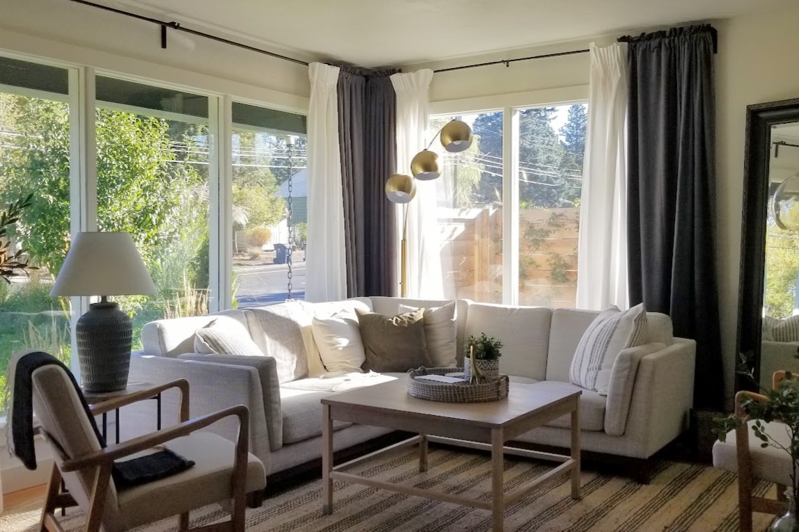 A spacious living area features large windows that allow natural light to fill the room. A comfortable sectional sofa is complemented by a coffee table, and a nearby lamp adds soft illumination. Neutral tones are accented by greenery visible outside.