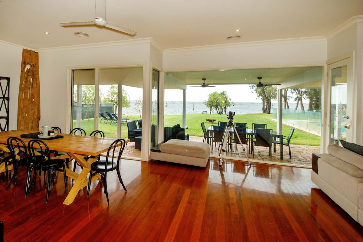 A spacious open-plan living area features a dining table with black chairs and a comfortable sofa. Large glass doors provide a view of the manicured lawn and water frontage, allowing natural light to fill the space. Ceiling fans are installed for air circulation.