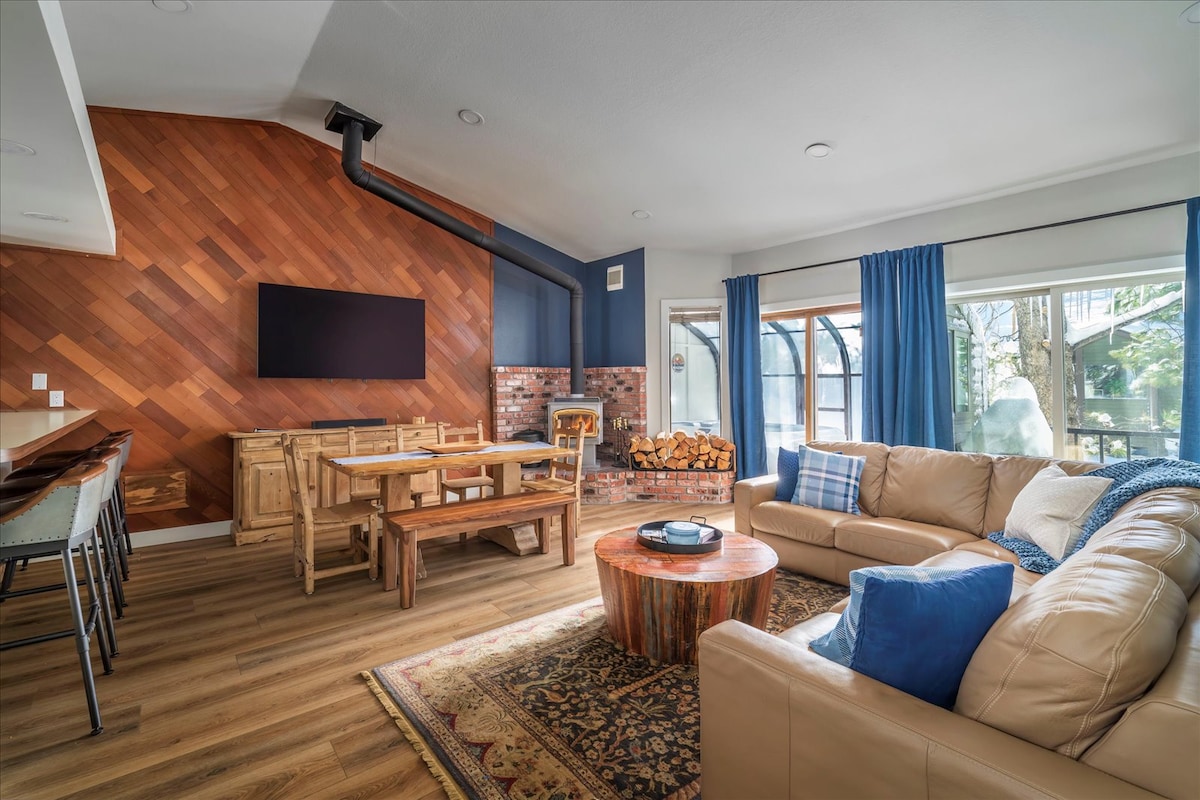 A spacious great room features a large sectional sofa and a rustic wooden coffee table. A dining table accommodates several guests, with a wood stove positioned beside a brick backdrop. Tall windows framed by blue curtains provide natural light and views of the surrounding trees.