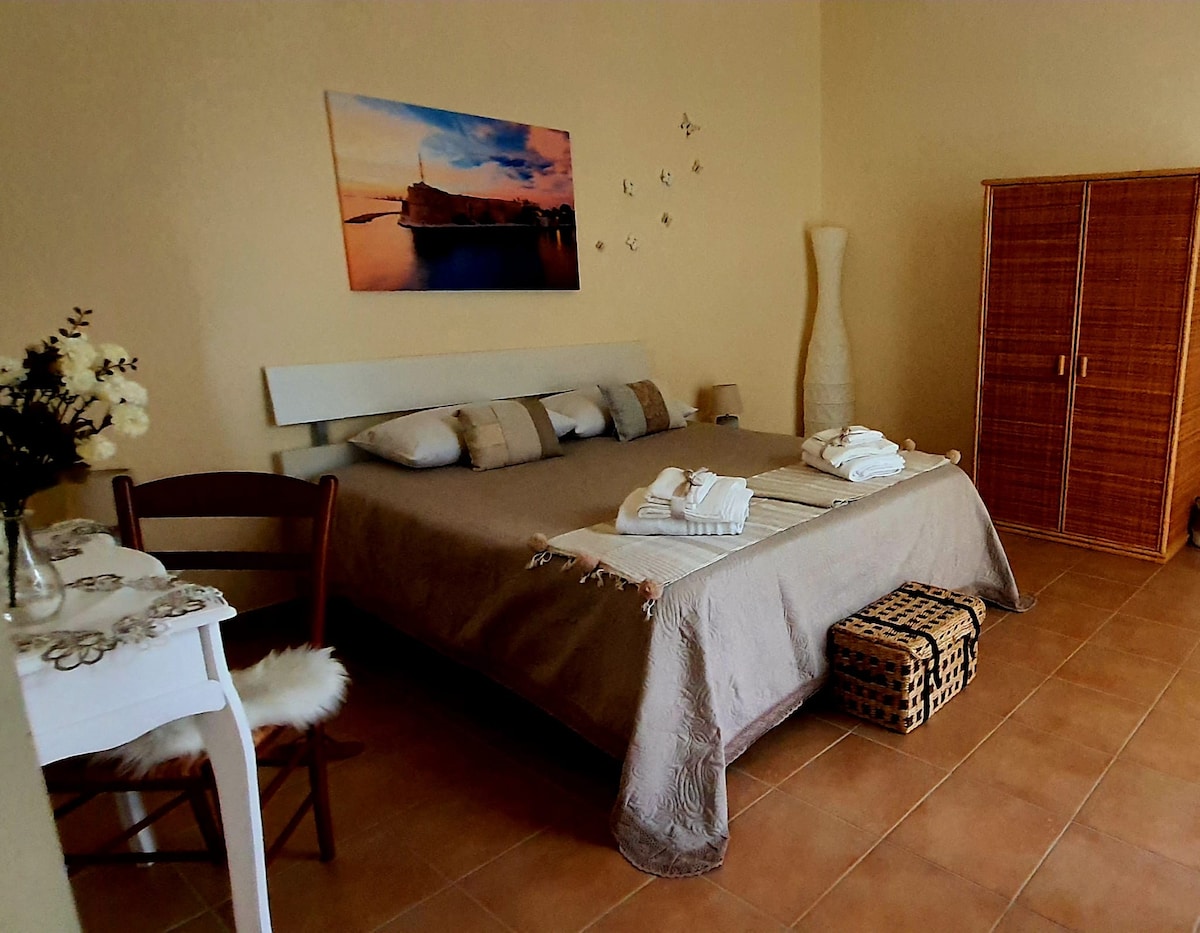 A spacious bedroom is furnished with a large bed covered in neutral bedding, accompanied by decorative pillows. A wall art piece depicting a city view hangs above the bed. A small table with a chair and an elegant lamp are positioned nearby, completing the inviting setting.