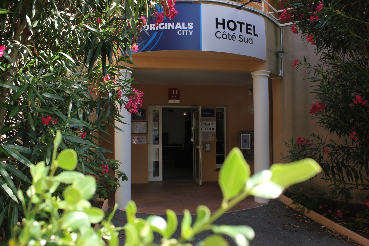 The hotel entrance is framed by lush greenery and vibrant flowers, creating a welcoming environment. A covered porch leads to the main door, marked with a clear sign displaying the hotel's name. The inviting exterior suggests a comfortable and accessible accommodation experience.