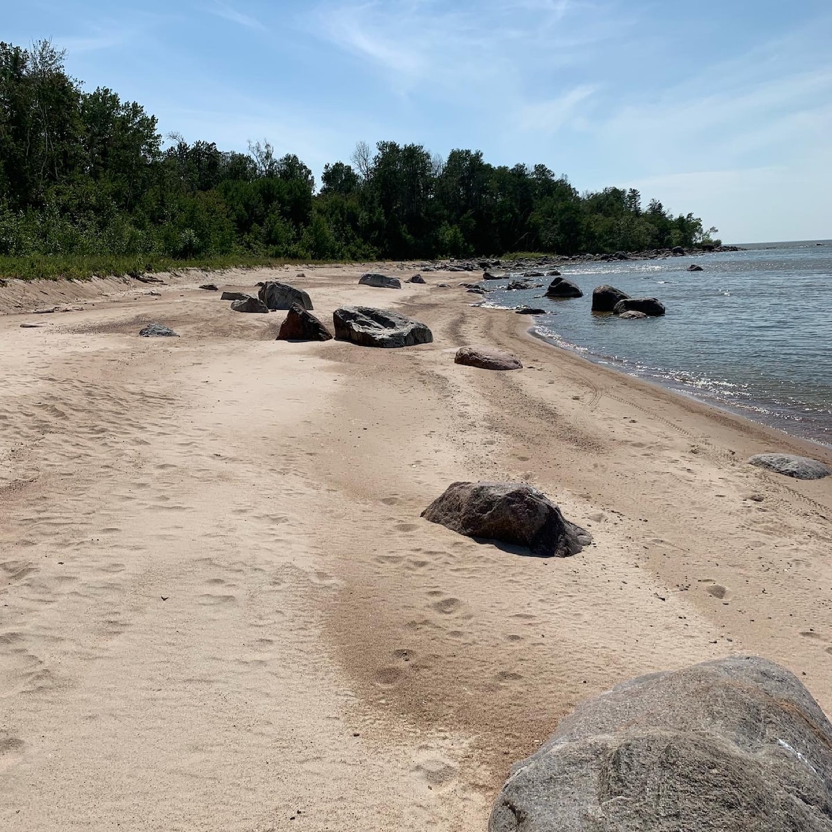 An expansive sandy beach stretches along the shoreline, framed by gentle waves. Smooth stones and boulders punctuate the landscape, blending with lush greenery in the background. The beach appears spacious, offering a serene environment for visitors seeking relaxation by the water.