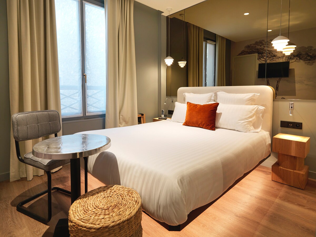 A modern bedroom features a spacious bed with crisp linens and an accent pillow. A small round table and a textured chair are positioned near a window dressed with soft curtains. Decorative lighting fixtures add a cozy touch to the room's neutral color palette.