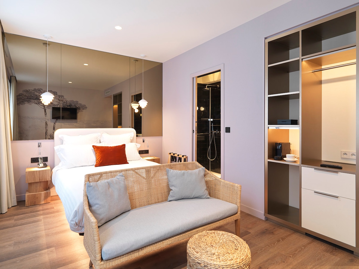 A spacious room features a double bed with a decorative pillow, a woven seating area, and a small round table. A modern shower is visible through a glass enclosure, and the room includes a built-in wardrobe with open shelving. Natural light enhances the contemporary decor.