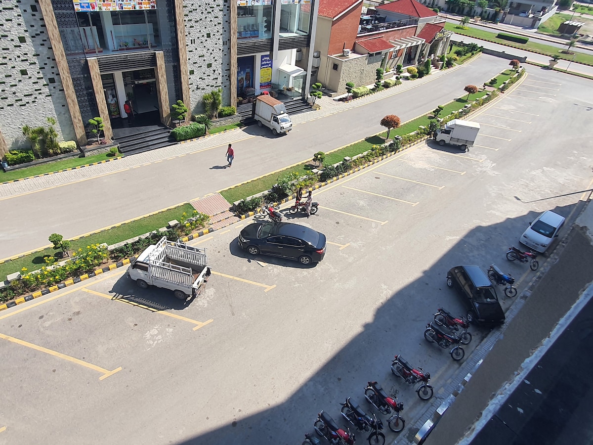A wide parking area accommodates multiple vehicles, including a black car and various motorcycles. Lush green plants line the edge of the lot, contributing to a tidy and orderly atmosphere. The surrounding buildings indicate a structured environment suitable for guests.