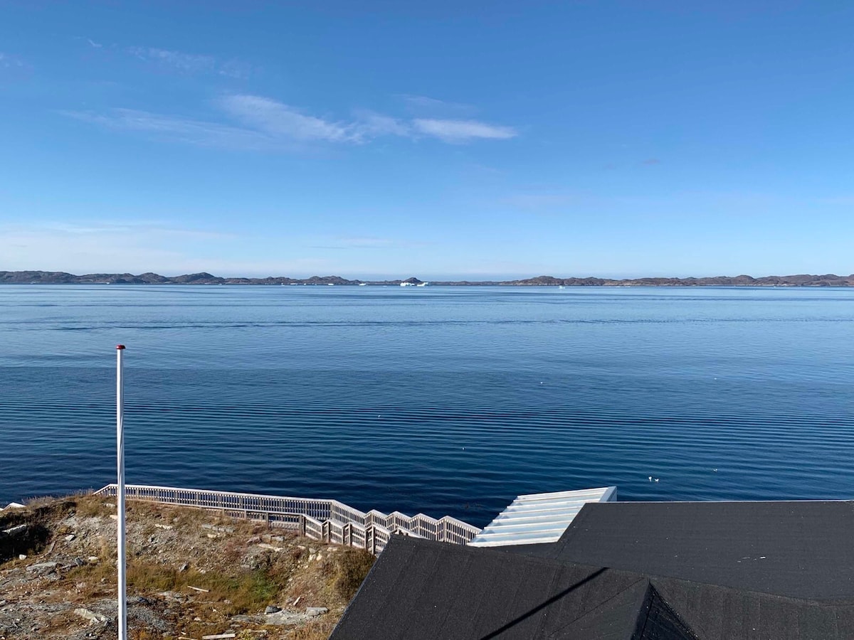 A serene coastal view is captured with calm waters reflecting the clear blue sky. Gentle waves lap against the shore, while distant landforms outline the horizon. A partially visible wooden structure extends along the waterfront, offering a natural setting for relaxation.