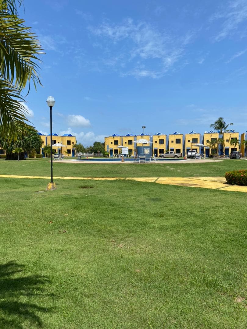 A well-maintained green lawn stretches across the foreground, leading to a collection of yellow buildings in the background. Pathways and palm trees are visible, contributing to the open and tranquil atmosphere of the vacation complex.