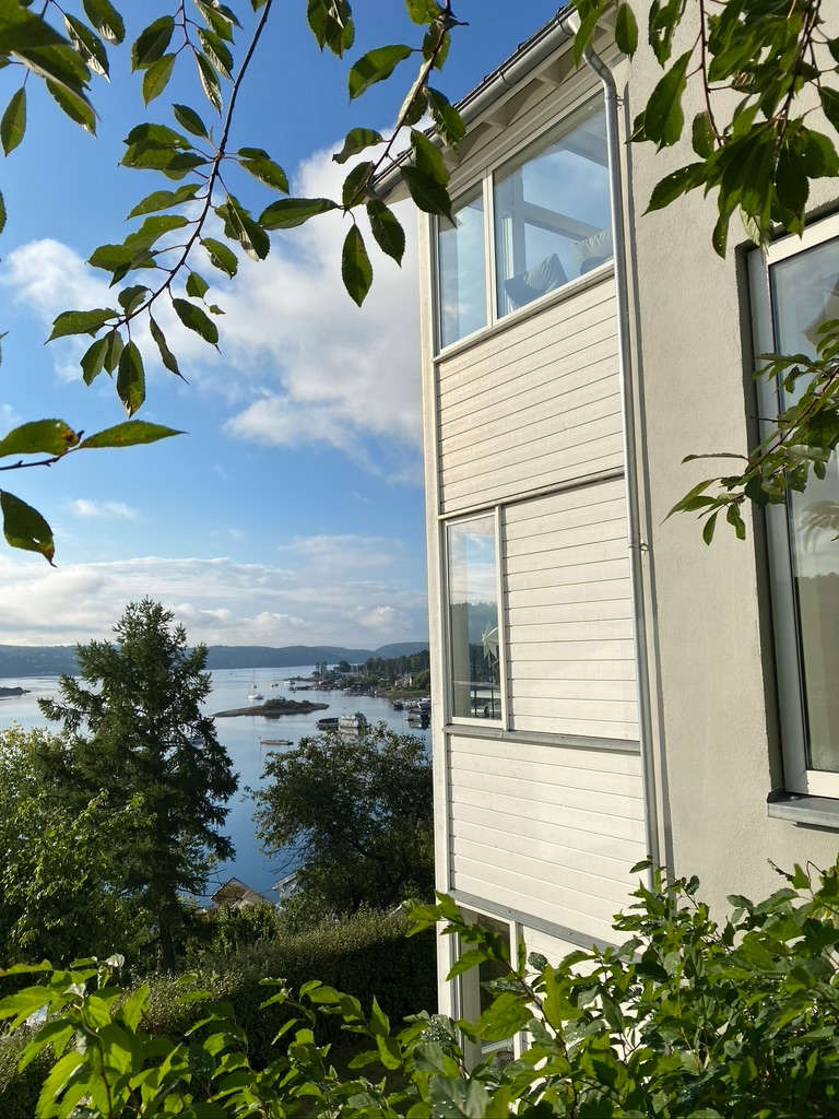 The exterior of a white house is framed by vibrant green leaves, showcasing large windows offering a view of the serene water and surrounding hills. The tranquil scene includes a glimpse of boats in the distance, enhancing the connection to the natural surroundings.