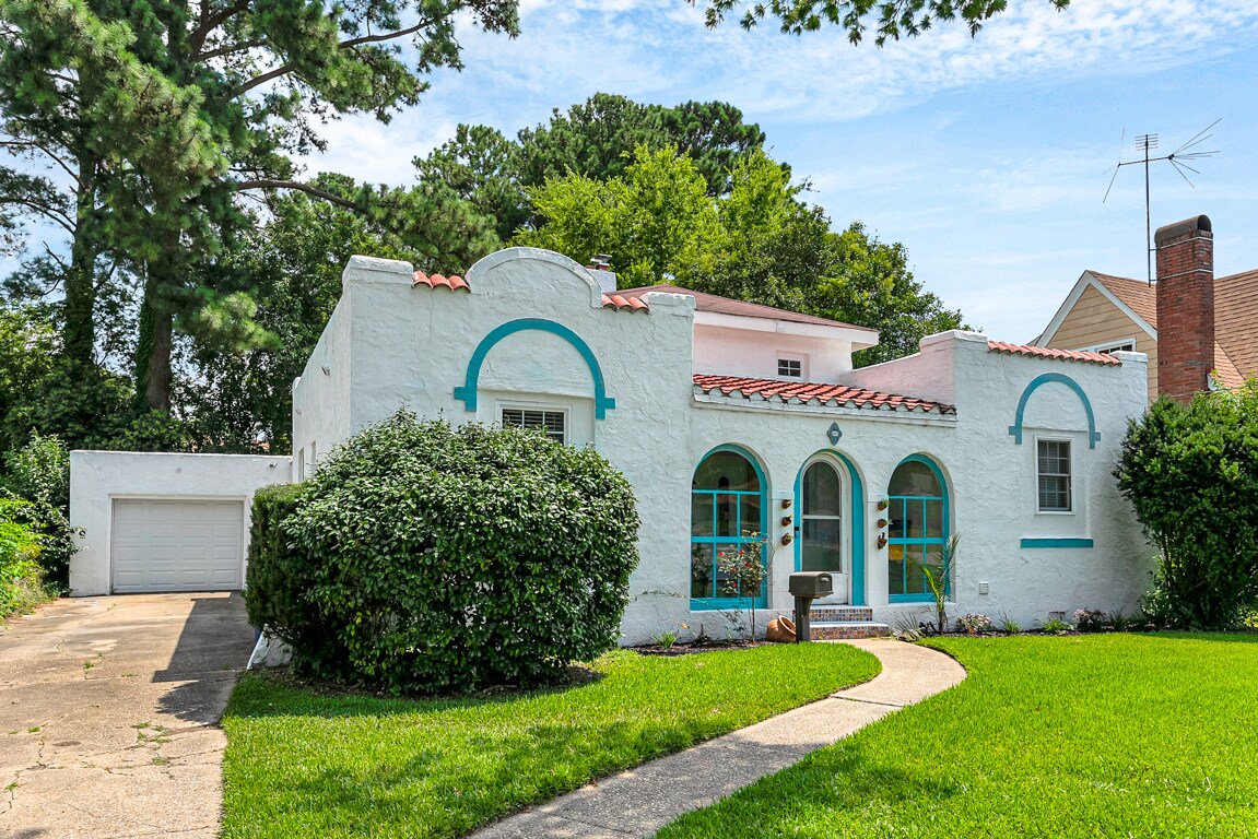 A charming white home with turquoise accents captures attention with its distinctive architectural style. Lush greenery surrounds the property, enhancing its curb appeal. A neatly paved walkway leads to the inviting entrance, while a driveway is visible, providing convenient access to the garage.