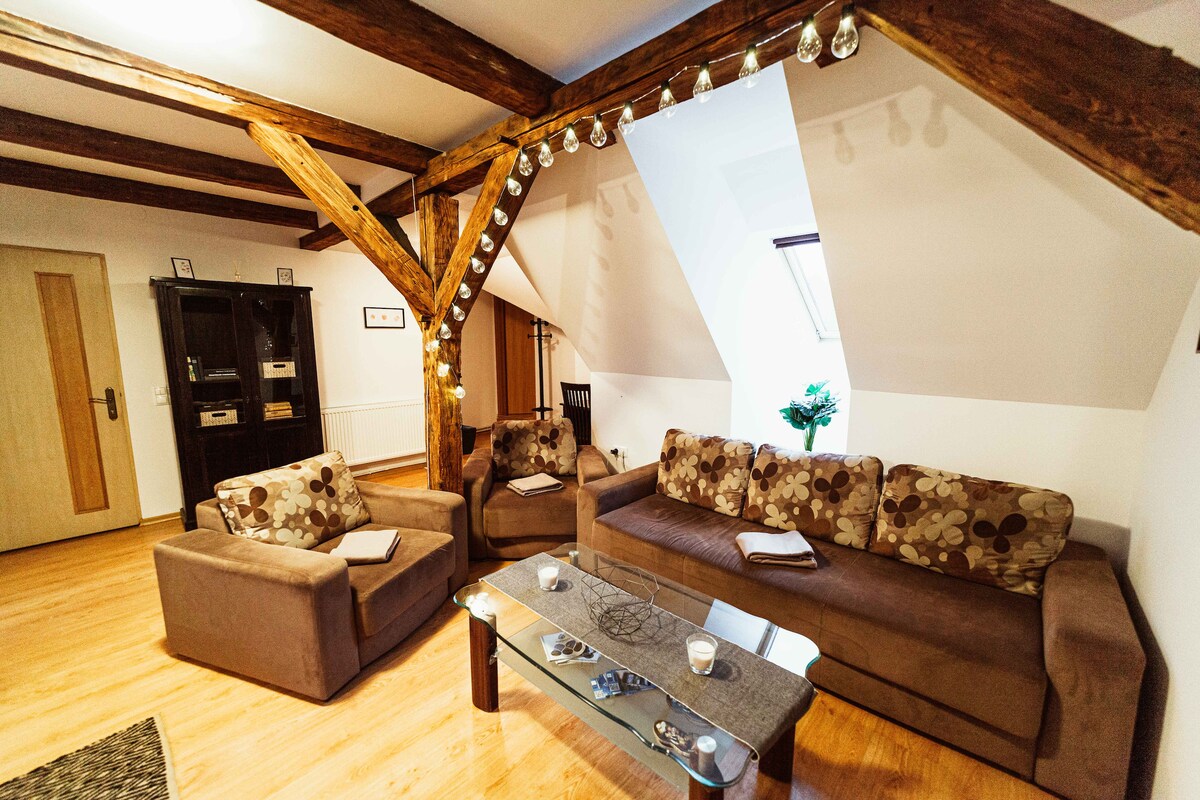 The living area is characterized by exposed wooden beams and a sloped ceiling. Two comfortable sofas are arranged around a glass coffee table, with decorative accents present. Natural light enters through a window, complemented by a small plant on a side table.