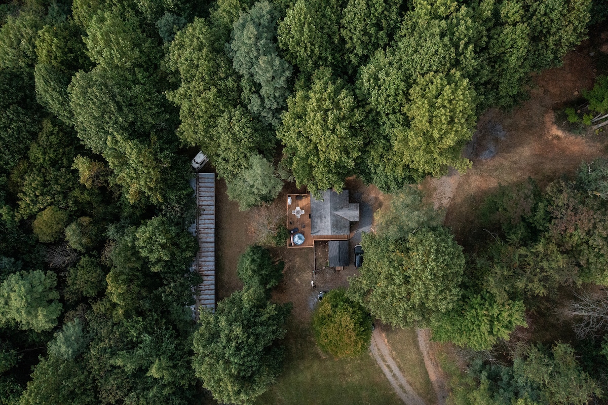An aerial view of a secluded cabin surrounded by dense greenery. The outdoor deck features a hot tub and seating area, offering a glimpse of the natural landscape. Paths meander through the trees, suggesting tranquility and privacy in a wooded setting.