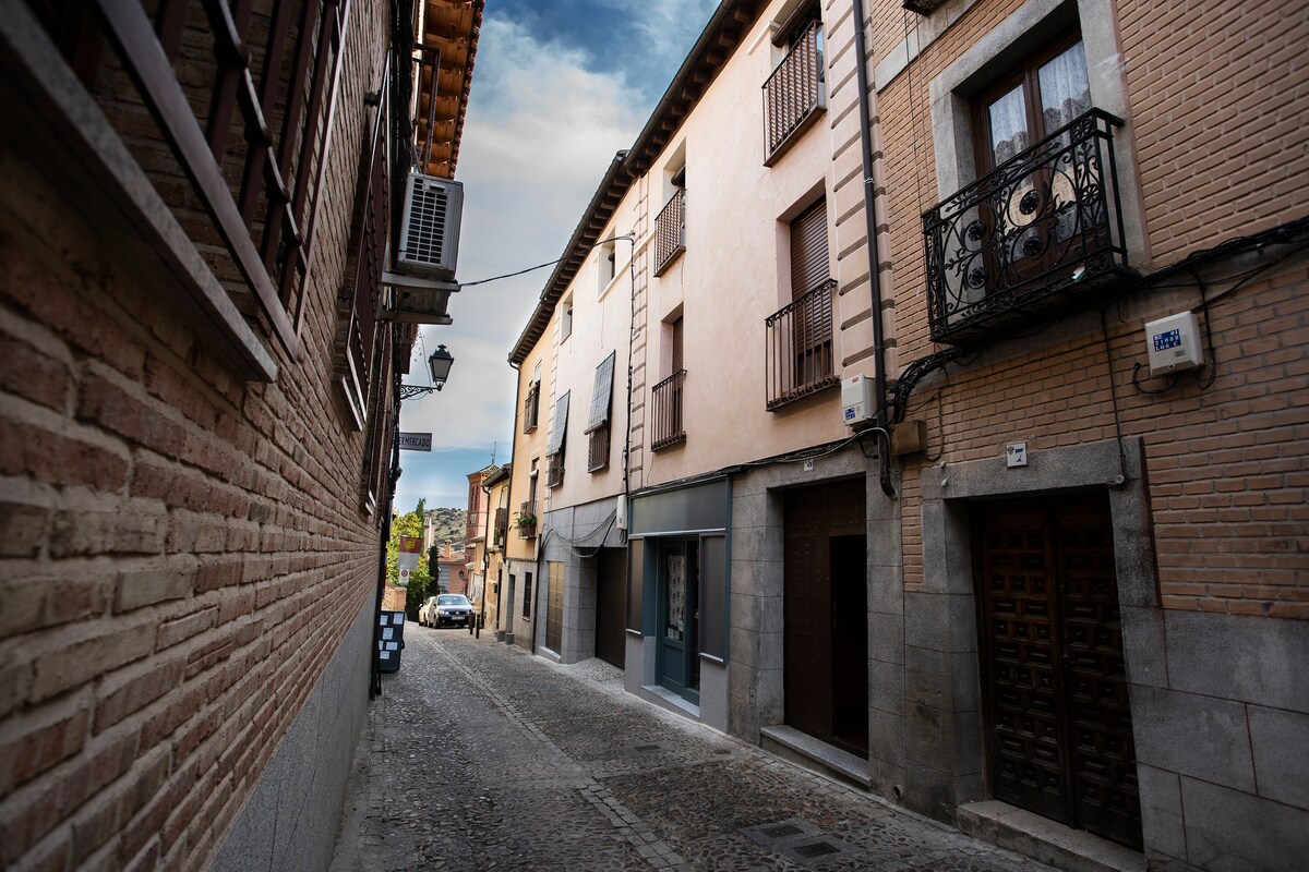 A cobblestone street framed by traditional buildings, featuring stone and brick façades. Balconies are visible, with a mix of modern and classic architecture. A parked car can be seen at the end of the street, and the sky is partly cloudy, providing a serene backdrop.