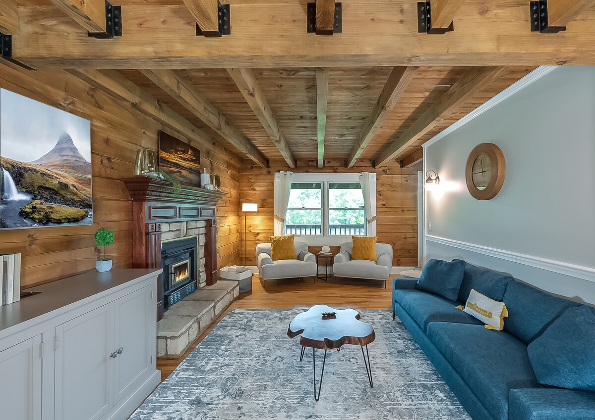 The living area features a wood-burning fireplace surrounded by stone, complemented by a large sectional sofa in rich blue. A round coffee table is positioned in the center, while natural light filters in through the window with a view of the outdoors.