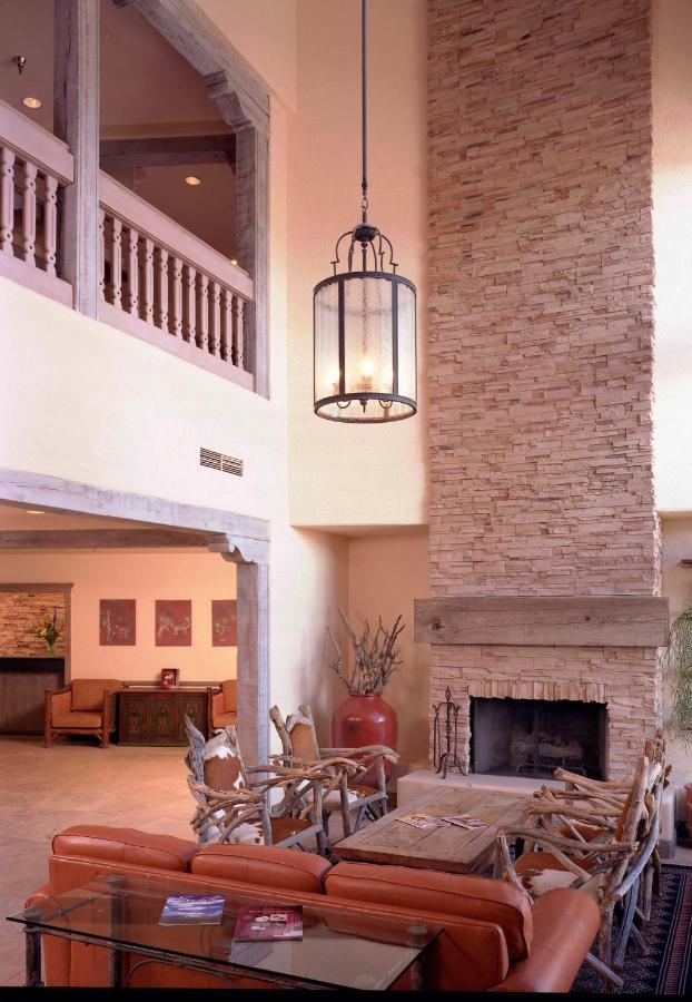 A spacious lobby area features a tall stone fireplace that serves as a focal point. Natural light highlights the warm tones of the wooden accents and rustic furniture. A large lantern hangs from the high ceiling, complementing the inviting atmosphere. Nearby reception area is subtly visible.