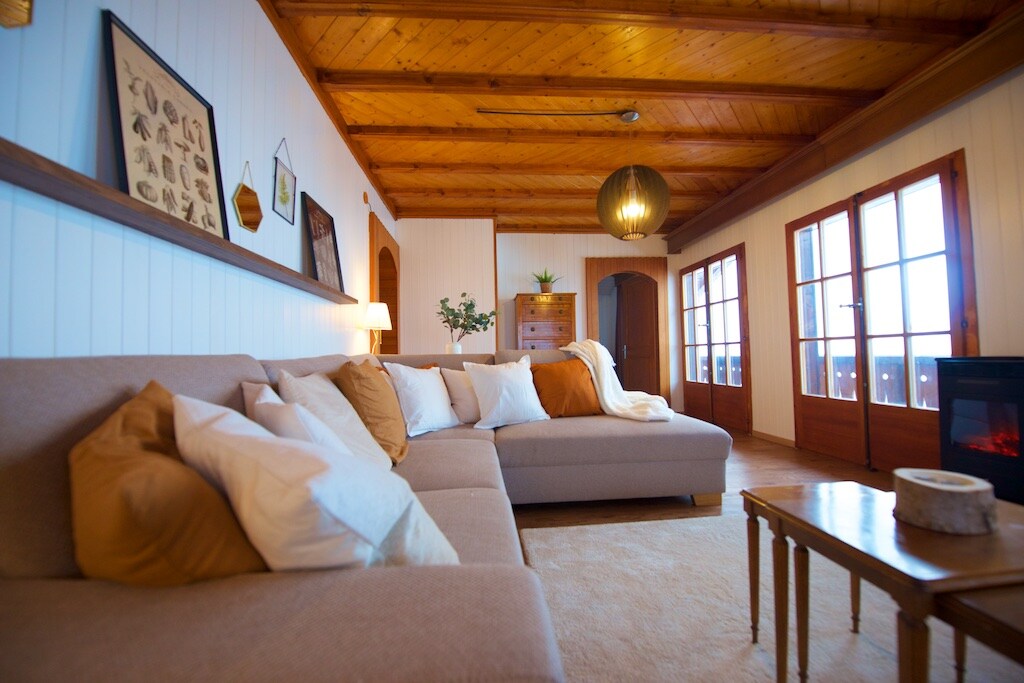 A spacious living area is highlighted by warm wood paneling and a high ceiling. A large, light-colored sectional sofa is arranged with multiple decorative pillows. Natural light enters through several windows, and a small wooden table enhances the cozy ambiance.