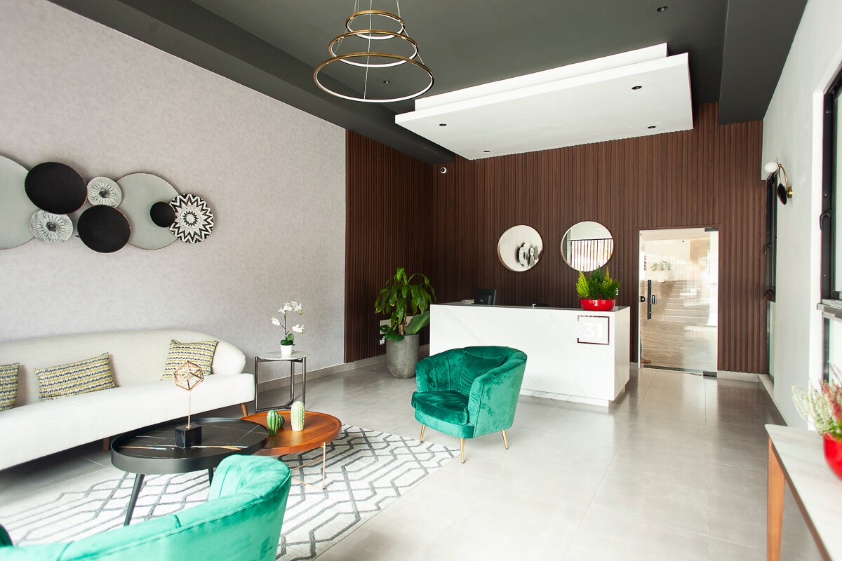 A lobby area features a modern design with a light-colored sofa and two green accent chairs. A wooden reception desk stands in the background, complemented by decorative wall art. Large windows allow natural light to illuminate the space, creating an inviting atmosphere.