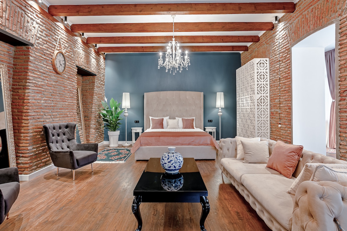 The studio features exposed brick walls and wooden beams, creating a spacious and inviting atmosphere. A king-size bed is centered in the room, complemented by a modern chandelier overhead. A Chesterfield sofa and lounge chairs provide comfortable seating around a dark coffee table.