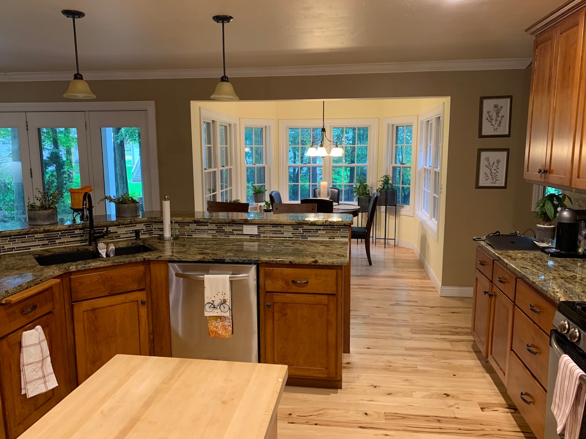 The spacious kitchen features wooden cabinets and granite countertops, complemented by modern appliances. A large island provides ample workspace. Through a set of glass doors, a charming dining area with multiple chairs and a sunny view of the backyard is visible.