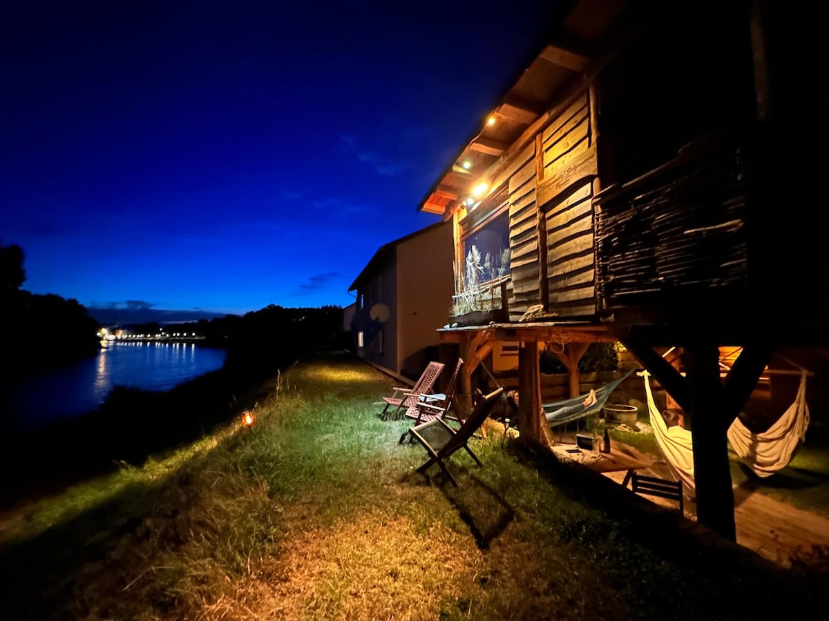 The image captures a wooden structure illuminated against a twilight sky. Relaxing seating options are provided, including lounge chairs and a hammock. A serene river is visible nearby, contributing to the tranquil atmosphere of the outdoor space.