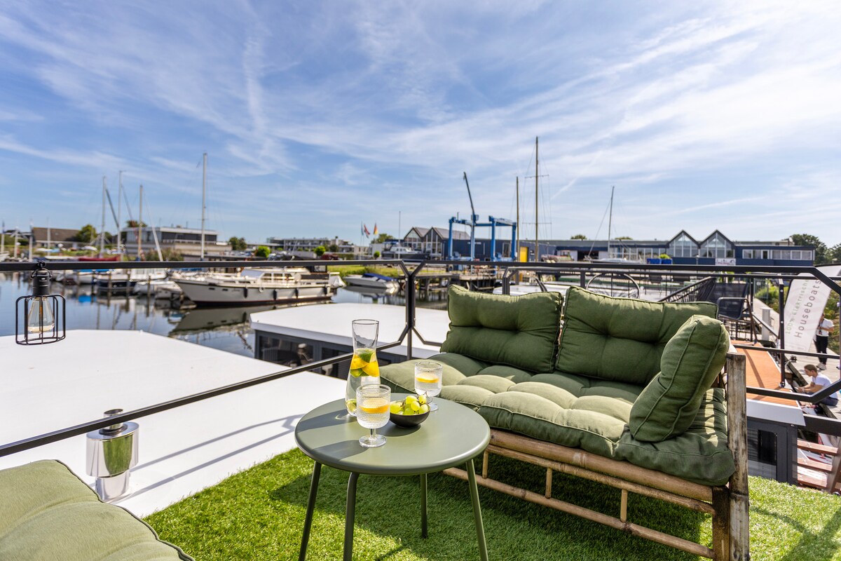 A comfortable outdoor seating area is presented on a spacious terrace, featuring a cozy green sofa and a small table. Refreshing drinks are served on the table, with the marina and its boats visible in the background under a clear blue sky.