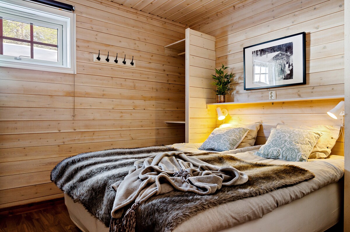 A comfortable bedroom features wood-paneled walls, creating a warm atmosphere. A spacious bed is layered with soft blankets and decorative pillows. A window brings natural light, and a wall-mounted photo adds an artistic touch. Shelving units and hooks provide practical storage options.
