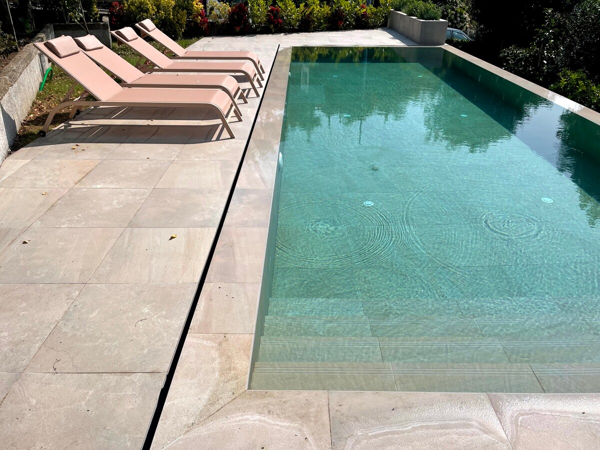 A sleek swimming pool reflects the surrounding greenery, bordered by smooth, light-colored stone. Five lounge chairs in soft pink are arranged nearby, providing a serene area for relaxation. Gentle ripples in the water suggest a calm atmosphere, inviting guests to unwind.