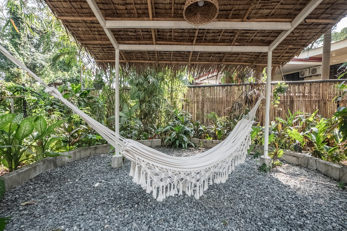 A shaded outdoor space is presented with a white hammock suspended beneath a thatched roof. Surrounding the hammock, lush greenery and tropical plants are visible, creating a tranquil and inviting atmosphere. Gravel covers the ground, providing a natural texture to the setting.