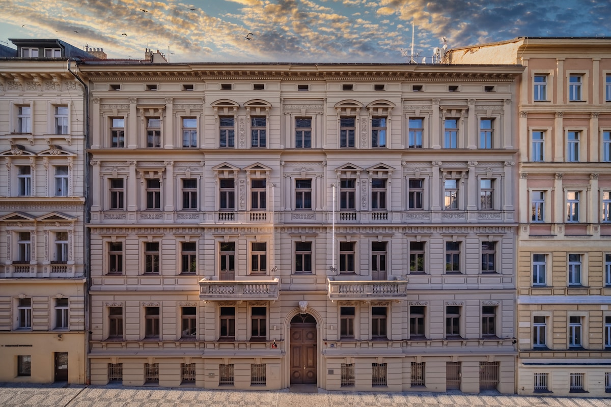 The historic building features a classic façade with elegant architectural details such as balconies and arched windows. The façade is adorned with soft colors, and the entrance is highlighted by a prominent doorway. The sky above reflects a mix of warm and cool tones during twilight.