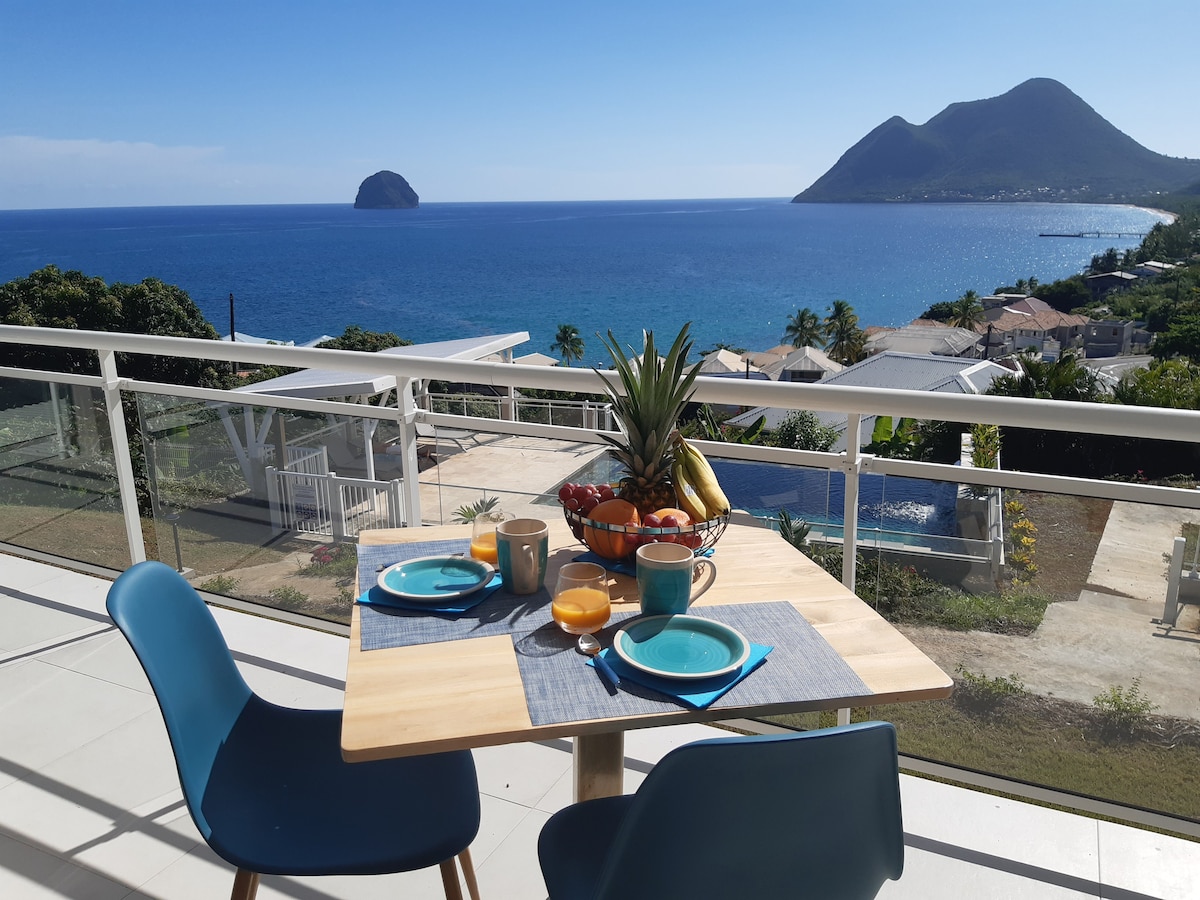 A dining table overlooking the Caribbean Sea is set with colorful plates and glasses. A bowl of fresh fruit, including bananas and oranges, is positioned at the center. The view features distant mountains and the sea, enhancing the outdoor dining experience.
