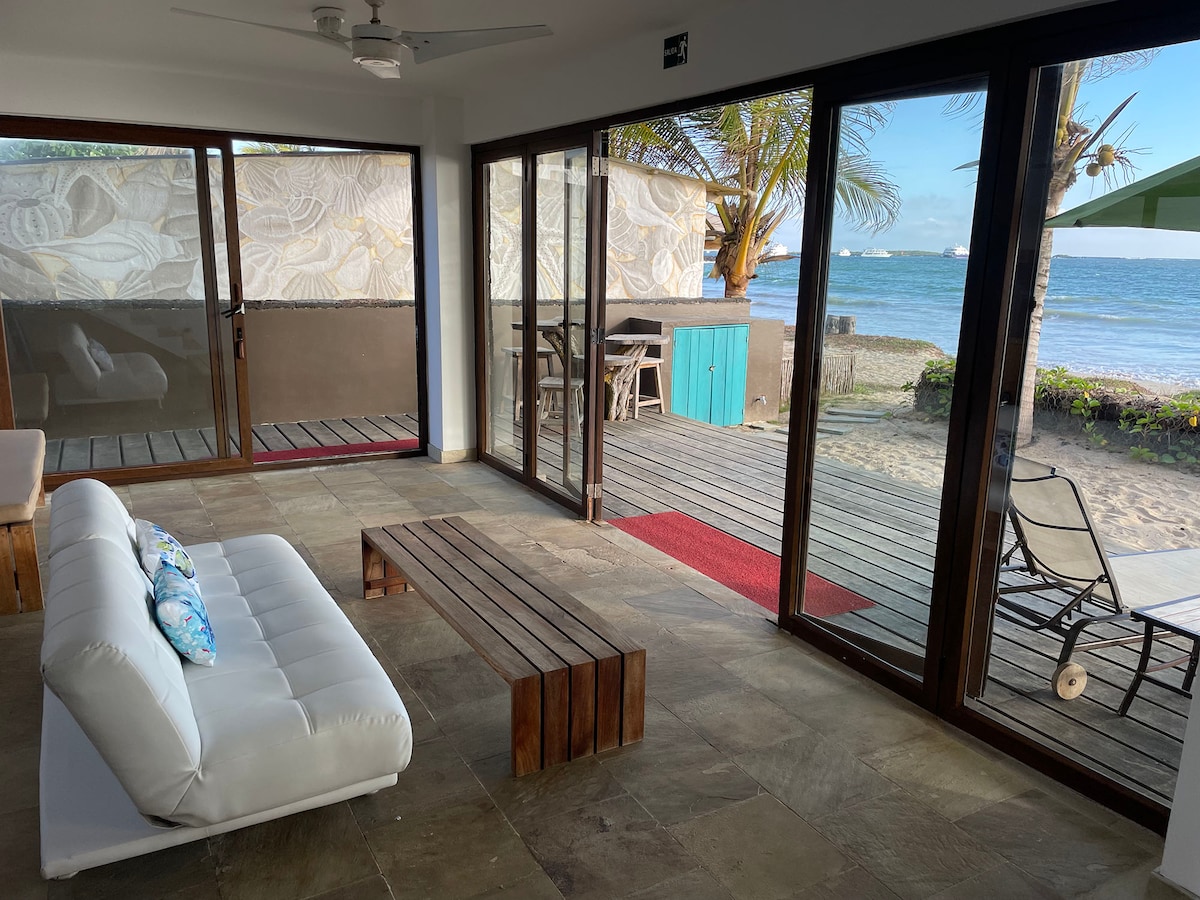 A welcoming living space features large glass doors that open directly to the beach, inviting natural light. Furnished with a minimalist white sofa and a wooden coffee table, the area connects seamlessly to the outdoor patio, where views of the ocean can be enjoyed.