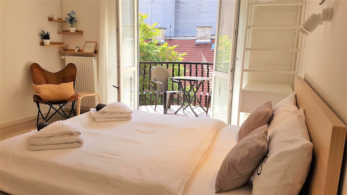 A light-filled studio apartment features a spacious bed with simple white linens and two decorative pillows. A small balcony overlooks greenery and includes a chair and table. A comfortable chair sits beside the bed, against a neutral wall with minimal decor.
