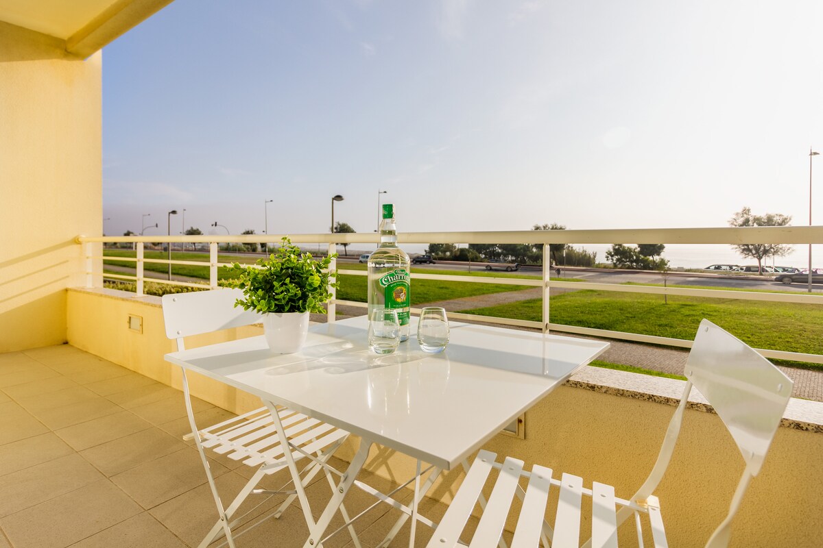 A serene balcony area is presented, featuring a small white table and two folding chairs. A potted plant adds a touch of greenery to the space. A panoramic view of the ocean and surrounding area is visible, enhancing the outdoor experience.