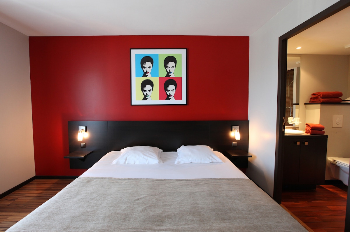 A spacious sleeping area features a dark wood bed frame with two white pillows and a light grey blanket. The wall behind the bed is painted a rich red, adorned with a colorful framed artwork depicting stylized faces. Soft lighting fixtures are positioned on either side of the bed.