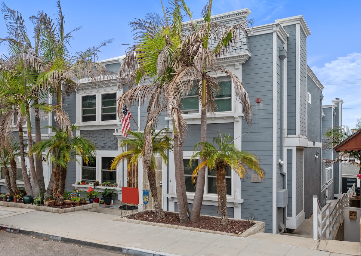 The exterior of a coastal home features light blue siding, adorned with palm trees and colorful flowers. An American flag is displayed prominently. A paved walkway leads to a white staircase, providing access to the entrance.