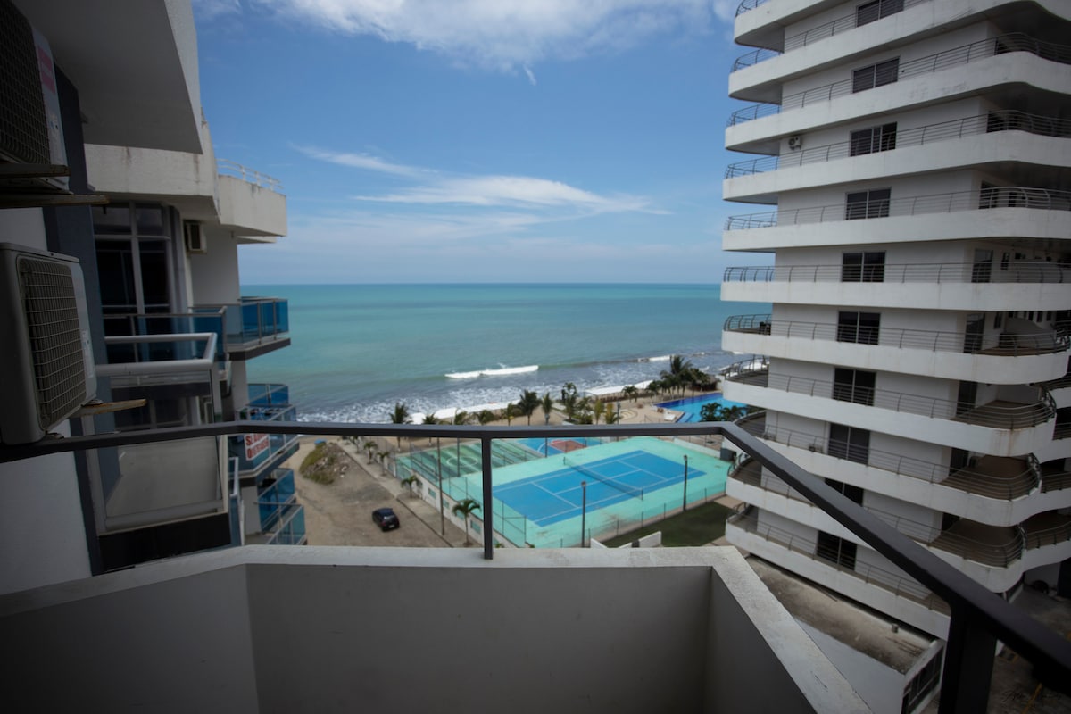 A balcony view offers a perspective of the ocean and beachfront. A turquoise sea meets the shore, while tennis courts with blue surfaces can be seen below. The buildings adjacent to the balcony create a contemporary coastal atmosphere with clear skies above.