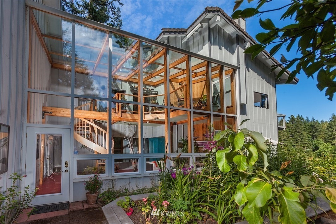 A sunlit exterior of the house is showcased, featuring a large solarium with expansive glass walls. A welcoming front entrance leads to a wooden staircase, while lush greenery and colorful flowers flank the pathway, complementing the home's natural surroundings.