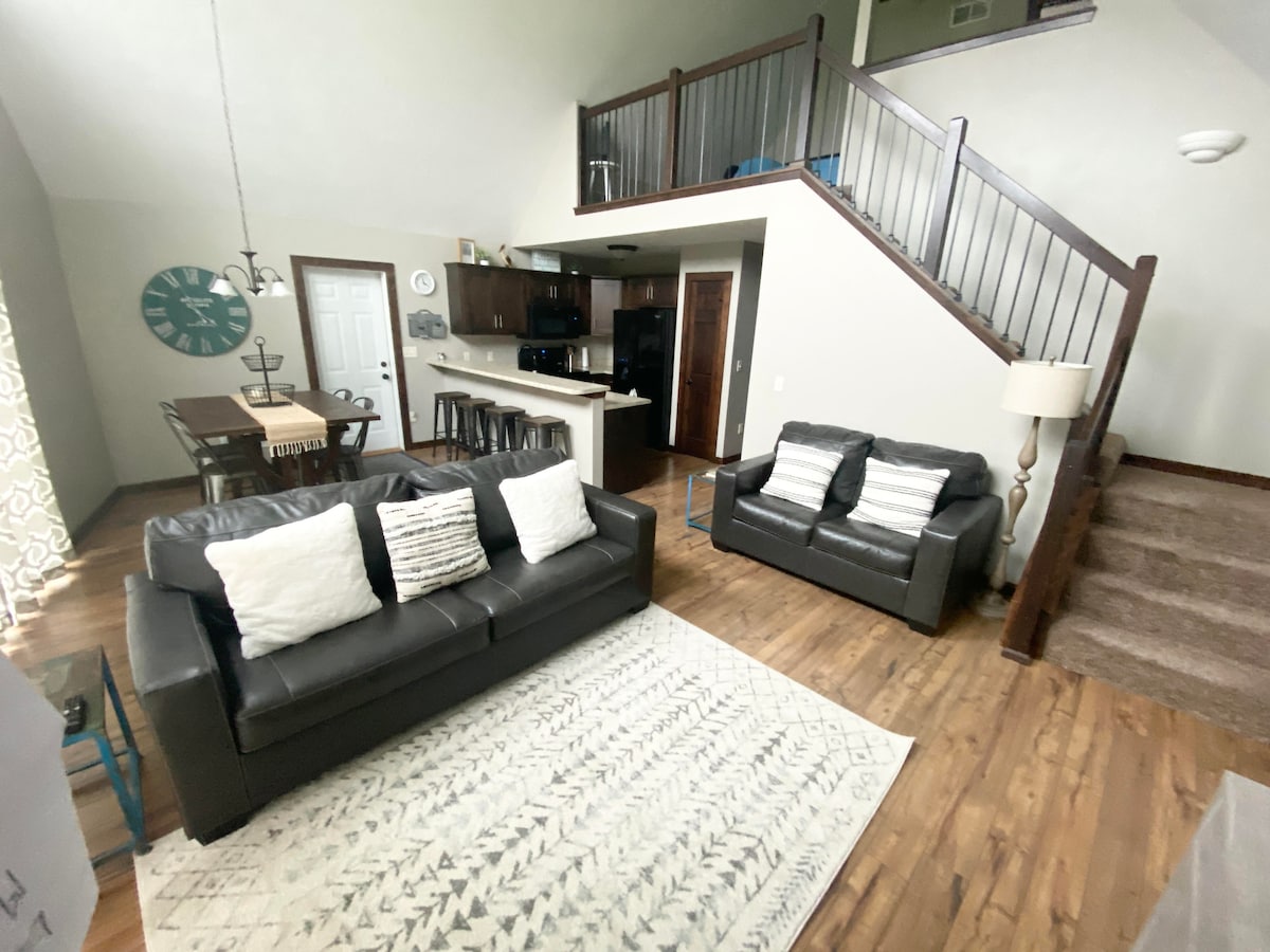 A spacious living area features a pair of leather sofas adorned with decorative pillows, set atop a light-colored area rug. A dining table is visible in the background, alongside a staircase leading to an upper level. Natural light enters through large windows covered with patterned curtains.