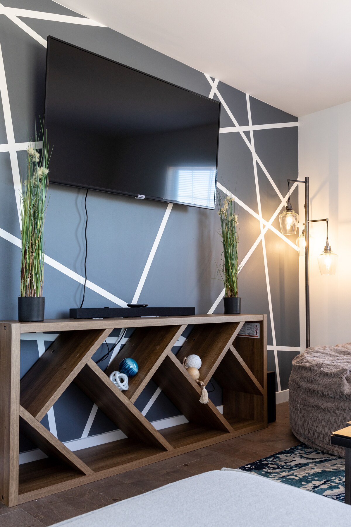 A modern living area features a large flat-screen TV mounted on a geometric patterned wall. Below, a wooden console table with an open design holds decorative items, while two potted plants add a touch of greenery. Soft lighting emanates from a nearby pendant lamp.