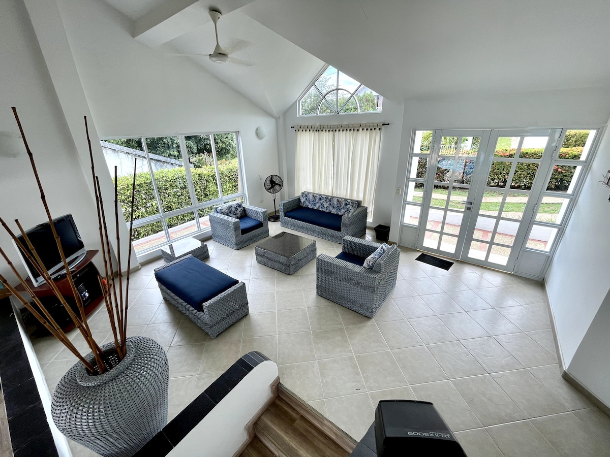 A spacious living area features large windows allowing natural light, with comfortable seating arranged around a central coffee table. A ceiling fan hangs overhead, and a television is positioned in the corner. The room opens to a view of the exterior greenery.