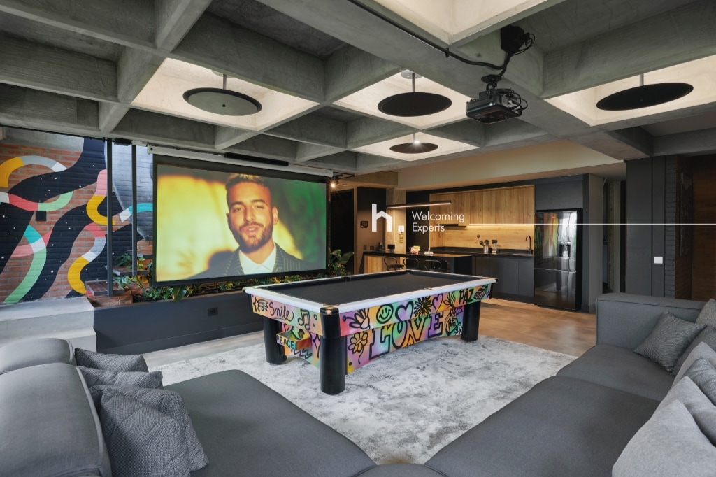A modern living space features a spacious sofa arranged around a colorful billiard table, adorned with graffiti art. A large screen is displayed in the background, complemented by vibrant mural decorations on adjacent walls. The kitchen is visible, showcasing sleek cabinetry.