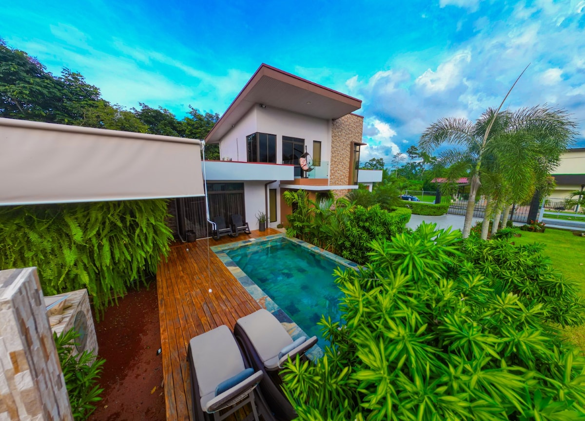 The exterior showcases a modern two-story house surrounded by lush greenery. A wooden deck leads to a clear blue pool, with two lounge chairs positioned nearby. The vibrant sky with scattered clouds complements the tranquil environment, creating an inviting outdoor space.