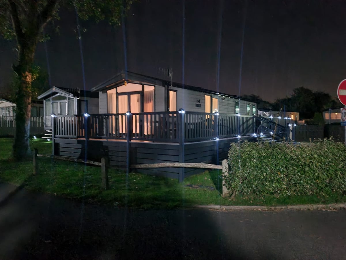 A lodge is situated prominently under night lighting, featuring a wrap-around veranda that allows access to the front and side of the building. The structure is flanked by a hedge and illuminated by soft outdoor lighting, enhancing its welcoming appearance.