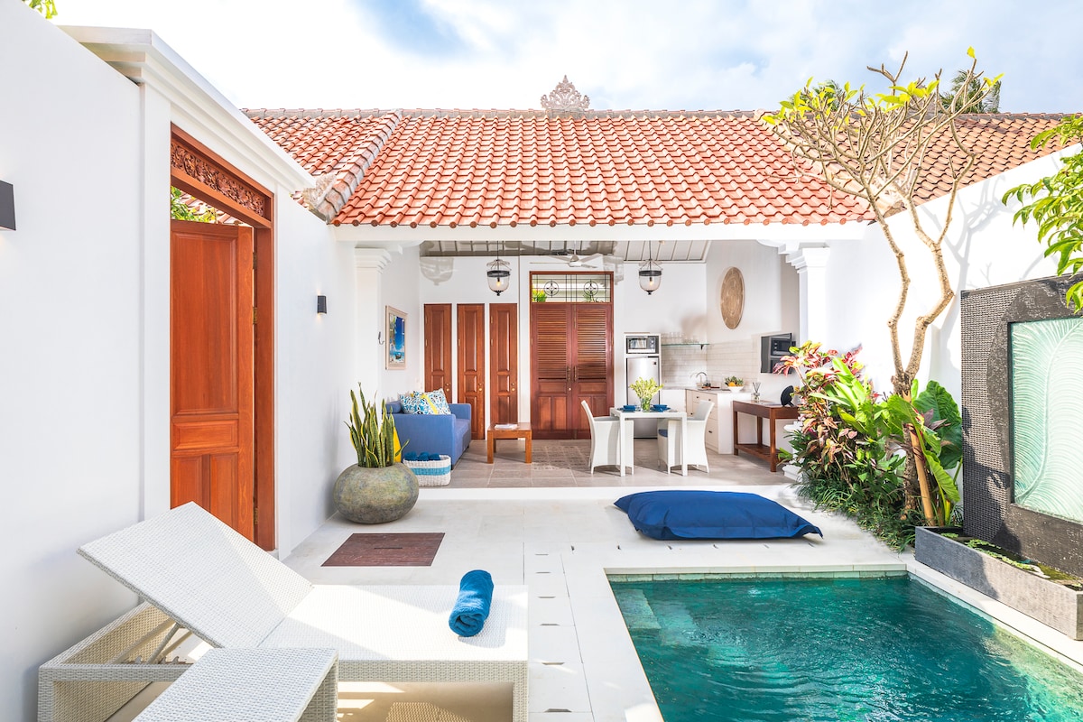 The villa's exterior is showcased with a sunlit private pool, surrounded by tropical plants. A cozy seating area is visible, featuring a blue armchair and a dining space with a fully equipped kitchen in the background. A calming water feature is situated beside the pool.