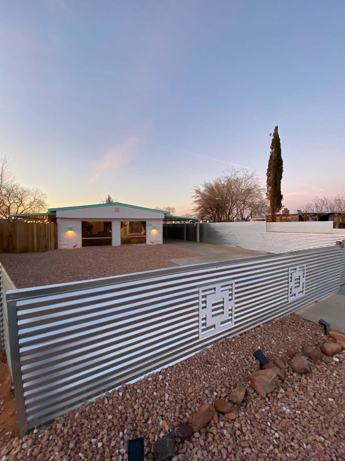A modern home is framed by a decorative corrugated metal fence, enclosing a gravel yard. Dusk light casts a soft glow on the house, highlighting the entryway and surrounding trees. The spacious outdoor area is ideal for relaxation and entertaining.