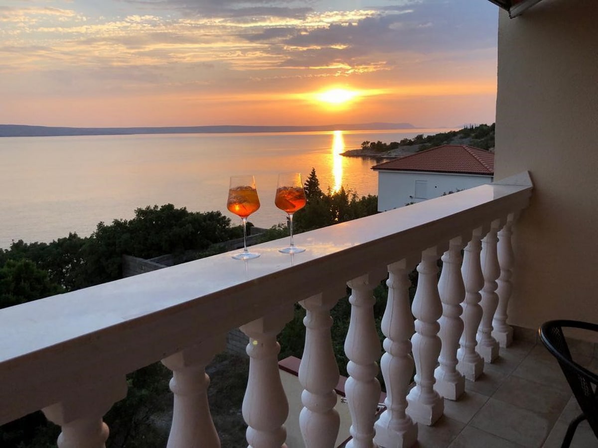A balcony railing with ornate balusters overlooks a calm sea at sunset. Two glasses filled with a beverage sit on the ledge, reflecting the warm hues of the sky. A gentle gradient of oranges, pinks, and purples is visible as the sun descends beyond the horizon.