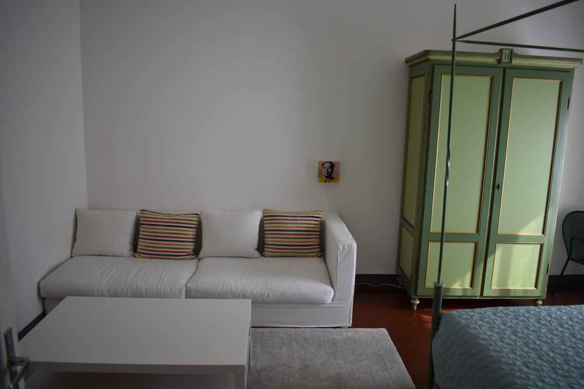 A bright room features a white sofa adorned with striped cushions, positioned beside a compact white table. An elegant green wardrobe stands against the wall, with a tastefully minimal art piece hanging nearby. Natural light reflects softly on the floor.