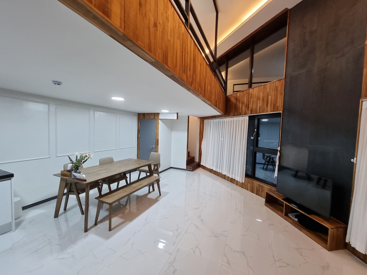 A dining area is featured with a long wooden table accompanied by light-colored chairs. Smooth white tiles reflect overhead lighting. A wooden accent wall adds warmth, while a large TV unit is positioned next to a staircase. Curtains frame large windows, allowing natural light to enter.